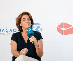 Mulher a falar com microfone, com logos da Fundação e da Praça da Fundacão ao fundo.
