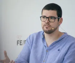 Homem de óculos, com camisa azul, a falar ao microfone. Fundo claro.