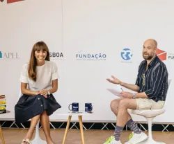 Duas pessoas a conversar em palco. Duas mesas, uma com livros e outra com canecas. Fundo com vários logotipos.