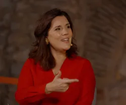 A advogada Assunção Cristas sentada, a falar. Uma parede de pedra como fundo.