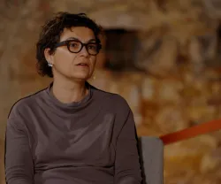 A economista Susana Peralta a falar, sentada numa cadeira. Parede de pedra ao fundo.