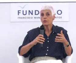 Uma mulher, de cabelo curto e branco, a falar num painel, com a inscrição "FUNDAÇÃO" ao fundo. Está a segurar um microfone.
