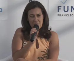Mulher a segurar num microfone, vestida com uma blusa amarela. Fundo branco com logotipo da Fundação.