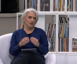 A jornalista Catarina Carvalho, sentada, a gesticular enquanto fala. Uma prateleira com livros ao fundo.