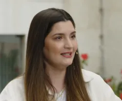 Mulher com cabelo longo e castanho, vestida com uma camisa branca, a falar.
