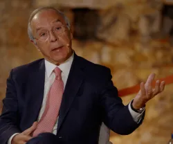 Homem (Luís Marques Mendes), a usar um fato azul e gravata rosa, sentado e a falar. Fundo de uma parede de pedra.
