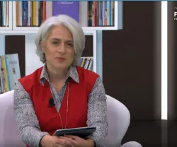Mulher de cabelo curto e cinza, vestida com um colete vermelho, sentada num sofá branco, com uma estante de livros atrás.