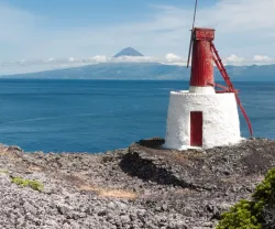Farol branco com detalhes vermelhos, à beira-mar, com o Pico ao fundo. Céu azul e nuvens. Créditos: Duarte Belo