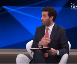 Carlos Oliveira Cruz sentado numa cadeira branca, a falar, enquanto segura um bloco de notas. Logótipo da Fundação no canto superior.
