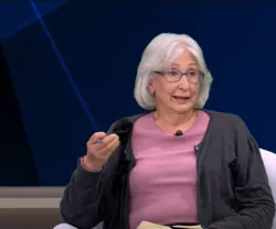 Mulher de cabelo grisalho, vestida com um casaco cinza e uma blusa rosa, a falar num estúdio de televisão.