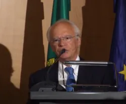 O professor Abel Mateus a falar ao microfone, com bandeiras ao fundo e copo de água à sua direita.