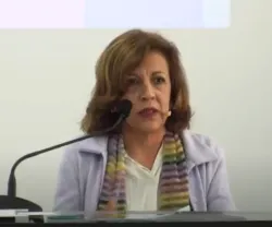 Mulher a falar num microfone, vestida com um casaco claro e lenço colorido. Fundo neutro.