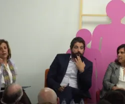 Três pessoas sentadas a discutir, com figuras humanas em rosa ao fundo.