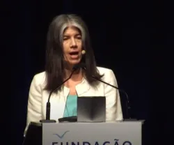 Mulher a falar ao microfone, com blazer branco e fundo escuro. Texto: "FUNDAÇÃO".