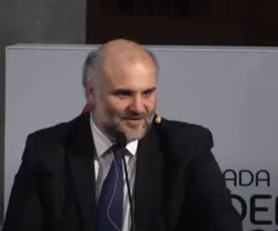 Homem de fato e gravata, a falar para um microfone. Fundo branco.