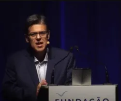 Homem com óculos fala ao microfone, com fundo azul e texto "FUNDAÇÃO" em destaque.