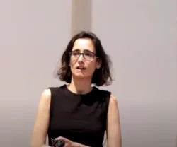 Mulher com óculos, vestida de preto, em palco, a falar para a audiência. Fundo claro.