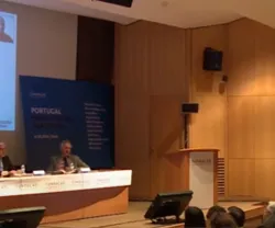 Imagem de uma conferência com painel de oradores e texto projetado atrás deles.