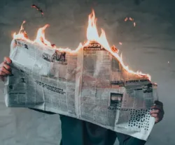 Pessoa segura um jornal em chamas contra um fundo cinza. Fogo na parte superior do jornal.