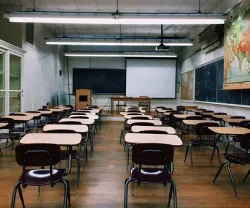 Sala de aula vazia, com mesas e cadeiras dispostas, quadro negro e mapa na parede.