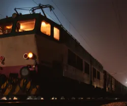 Locomotiva à noite com luzes acesas, transporte de vagões visível ao fundo.
