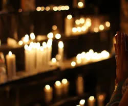 Mãos juntas em oração, diante de velas acesas, num espaço religioso.