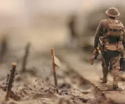 Soldado em miniatura caminha por um terreno de guerra, com mochila e arma, em foco.