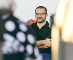 Homem com óculos, sorrindo, sentado, com uma camisa preta, a surgir ao fundo, no meio de duas pessoas, que estão de costas. 