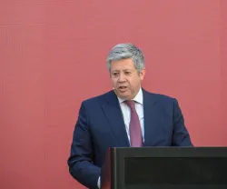 José Soares dos Santos, vestido de fato escuro, camisa branca e gravata vermelha, a discursar. Fundo rosa.