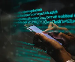 Mão a segurar num telemóvel com um código de programação no fundo.