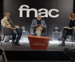 Três pessoas sentadas em cadeiras, num palco, durante um evento. Logo da FNAC ao fundo.