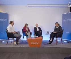 Painel de discussão com quatro participantes. Uma mesa laranja no centro do palco. Fundo branco.