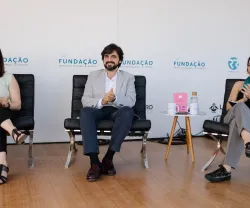Três pessoas sentadas em cadeiras, aplaudindo, com logotipos da FUNDAÇÃO ao fundo.
