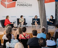 Uma plateia a assistir a um debate com quatro oradores em palco. Ao fundo, o texto "Praça da Fundação".