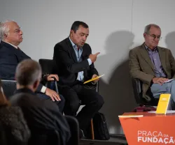 Três homens sentados em cadeiras, um deles gesticula. Texto: "PRAÇA DA FUNDAÇÃO" em destaque numa mesa laranja.