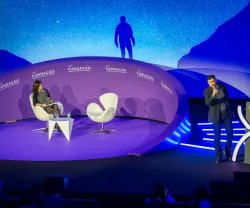 Imagem de um palco bem iluminado, com duas cadeiras brancas, um apresentador e texto "FUNDAÇÃO" ao fundo.