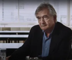 Homem de cabelo grisalho, Antony Beevor, sentado numa mesa vazia, copo de água ao lado. Ambiente escolar.