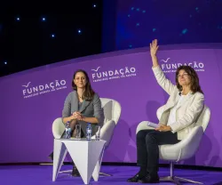 Duas mulheres sentadas, num palco com fundo roxo, uma delas está a acenar. Logo "FUNDAÇÃO".