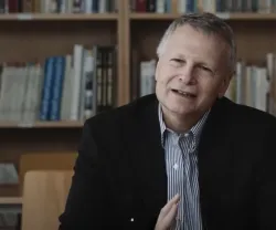 O economista político Dani Rodrik, de cabelo grisalho, sorrindo, sentado à mesa com prateleira de livros ao fundo.