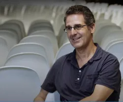 Mark Mazower, sorridente, sentado em frente a cadeiras brancas vazias. Sem texto na imagem. 