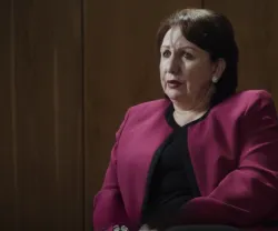 Mulher com blazer rosa sentado, expressão séria, num ambiente com paredes de madeira.