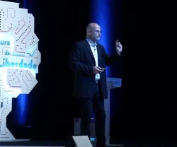 Yochai Benkler de pé num palco, gesticulando enquanto faz uma apresentação ao lado de um painel em forma de árvore com o texto "À procura da Liberdade".