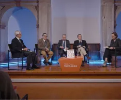 Imagem de um painel com cinco pessoas sentadas em cadeiras, com o texto "Fundação" visível numa mesa laranja que está ao centro.