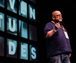 O sociólogo John Goodwin a falar em palco, a usar uma camisola escura e óculos. Texto visível: "juventudes".