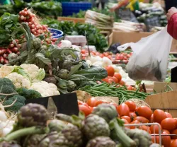 Imagem de mulher a comprar vegetais numa praça. Crédito: Canva