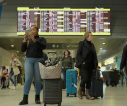Mulher a olhar para o telemóvel em frente a um painel de chegadas e partidas do aeroporto de Lisboa.