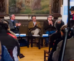 Evento com público a aplaudir, três oradores em destaque em frente a um quadro artístico.