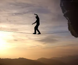 Imagem de um homem a caminhar numa arame suspenso entre duas rochas. Crédito: Canva