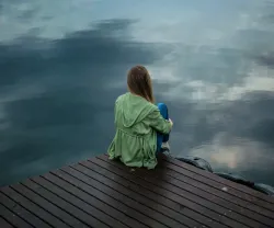 Imagem de uma mulher de costas, pensativa, junto a um lago. Crédito: Keenan Constance/Canva