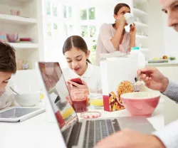 Uma família toma o pequeno-almoço na cozinha, mas o pai e os dois filhos estão a ver ecrãs de computador ou telemóvel. Crédito: Canva